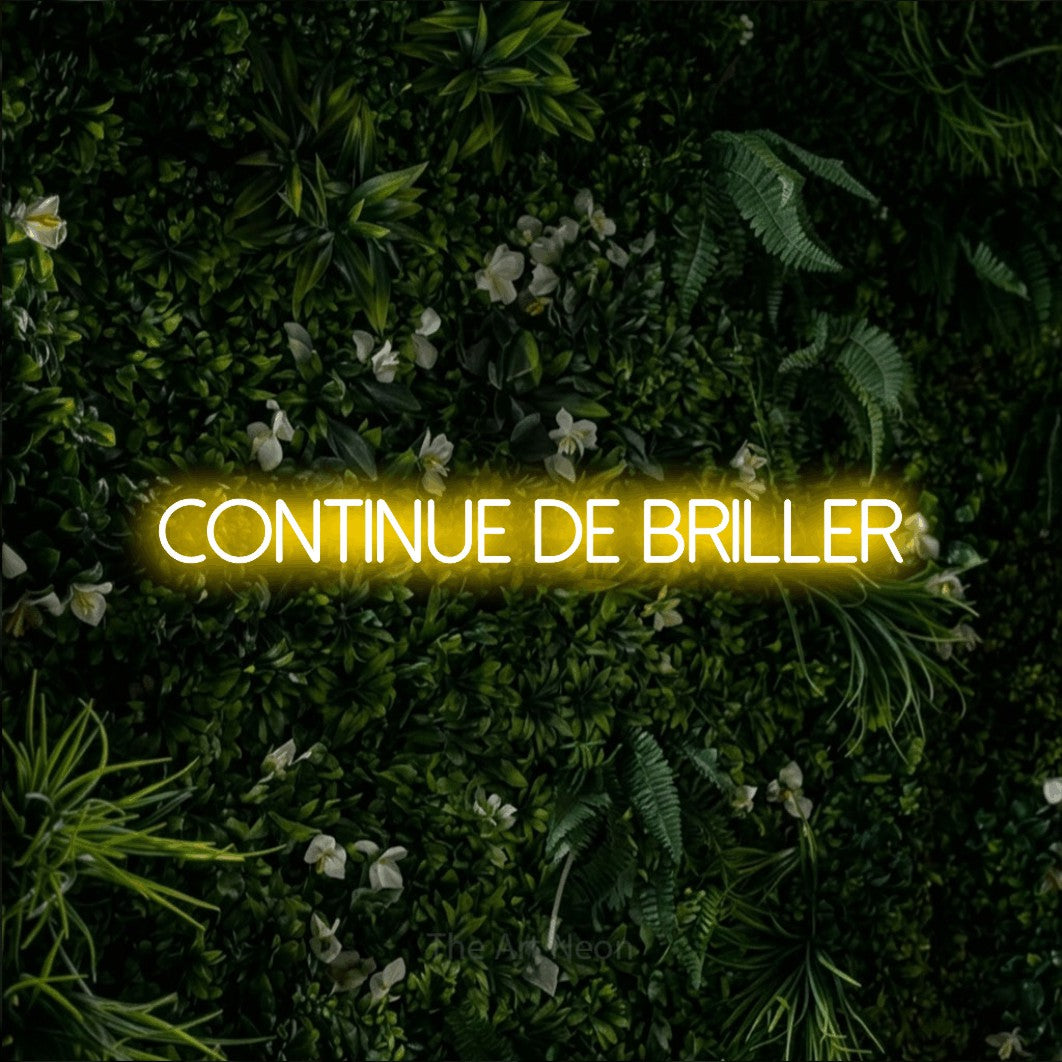 “Continue de briller” LED neon sign in yellow, with thin sans-serif lettering, shown against a dense green plant wall.