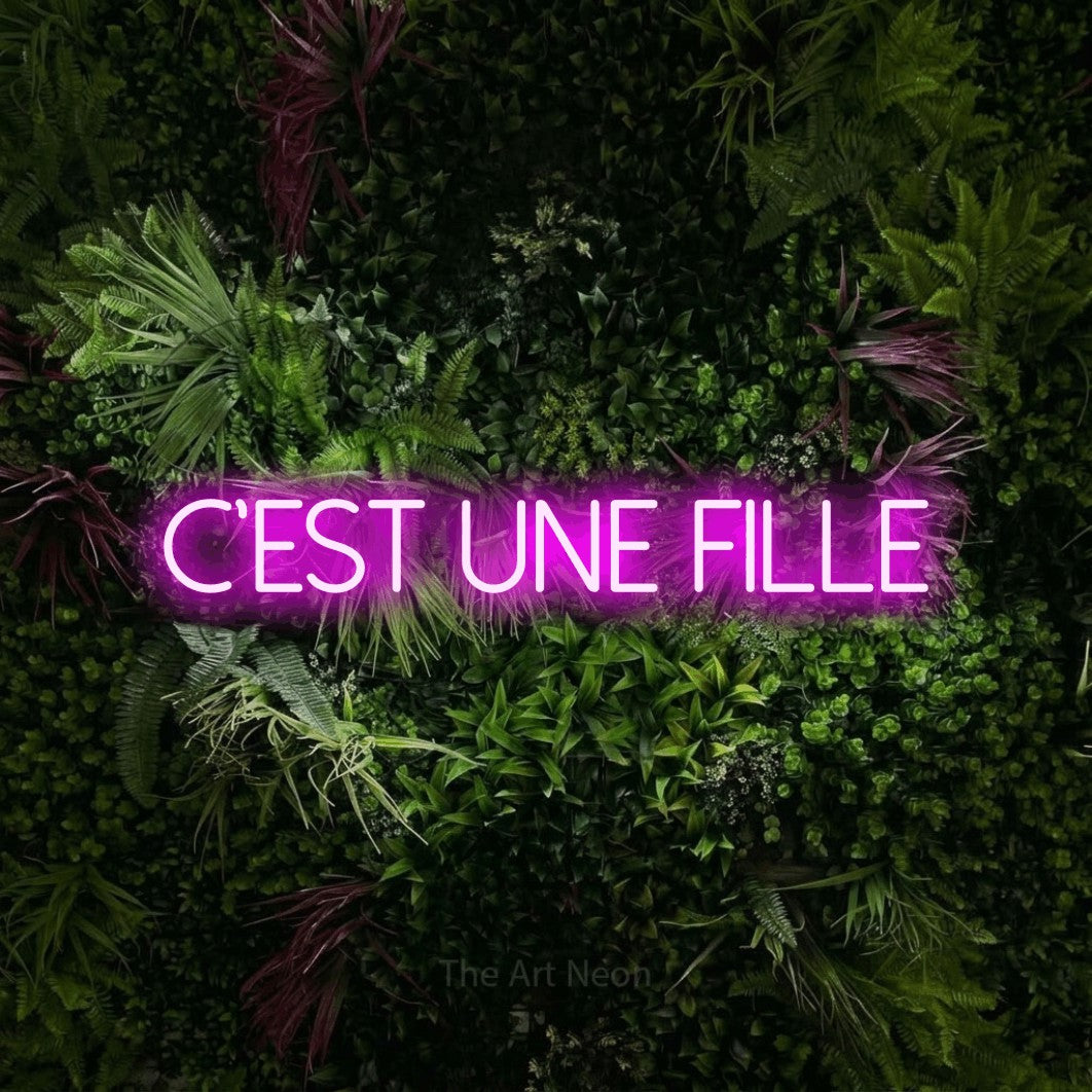 Néon LED « C'est une fille » en rose, en lettres fines sans empattement, mis en scène sur un mur végétal aux fleurs roses, pour une fête prénatale.