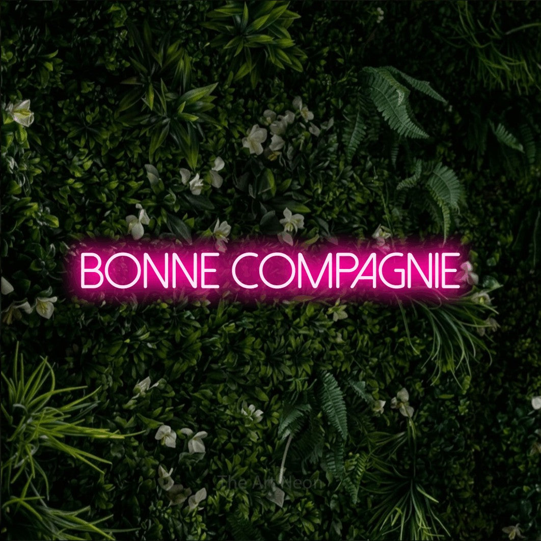 “Bonne compagnie” LED neon sign in hot pink, with thin sans-serif lettering, shown against a dense green plant wall.