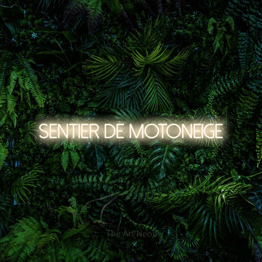 “Sentier de motoneige” LED neon sign in warm white, with thin sans-serif lettering, shown against dense tropical foliage.