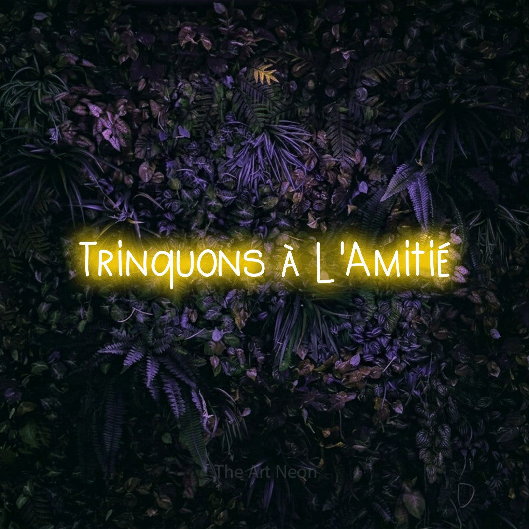 “Trinquons à l'amitié” LED neon sign in yellow, with narrow hand-drawn lettering, shown against a dark wall with purple flowers.