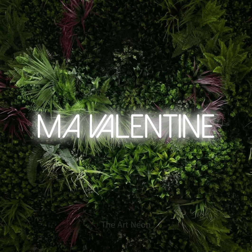“Ma valentine” LED neon sign in cool white, with thin sans-serif lettering, shown against a plant wall with pink flowers.