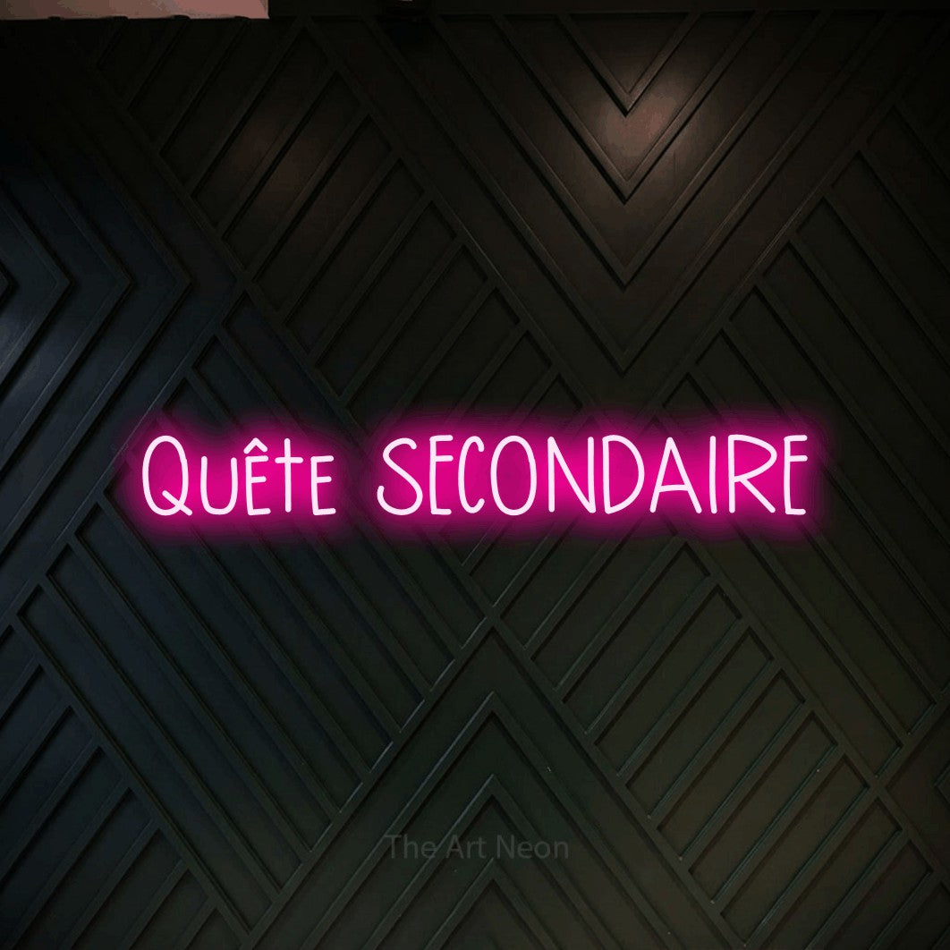“Quête secondaire” LED neon sign in hot pink, with narrow hand-drawn lettering, shown against a black chevron-patterned wood wall.