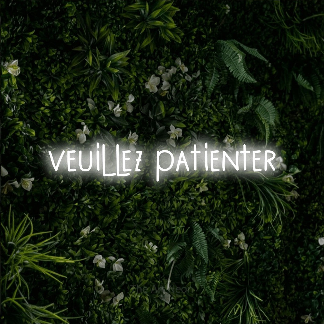 “Veuillez patienter” LED neon sign in cool white, with narrow hand-drawn lettering, shown against a dense green plant wall.