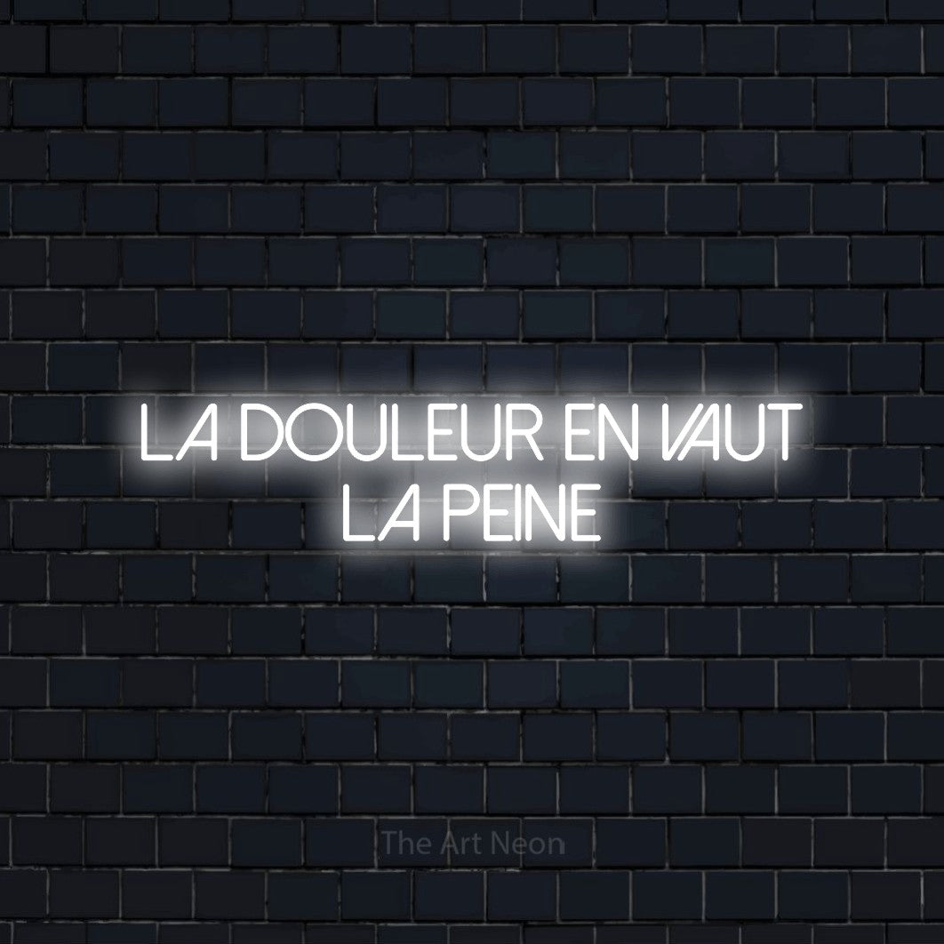 “La douleur en vaut la peine” LED neon sign in cool white, with thin sans-serif lettering across 2 lines, shown against a dark navy brick wall.