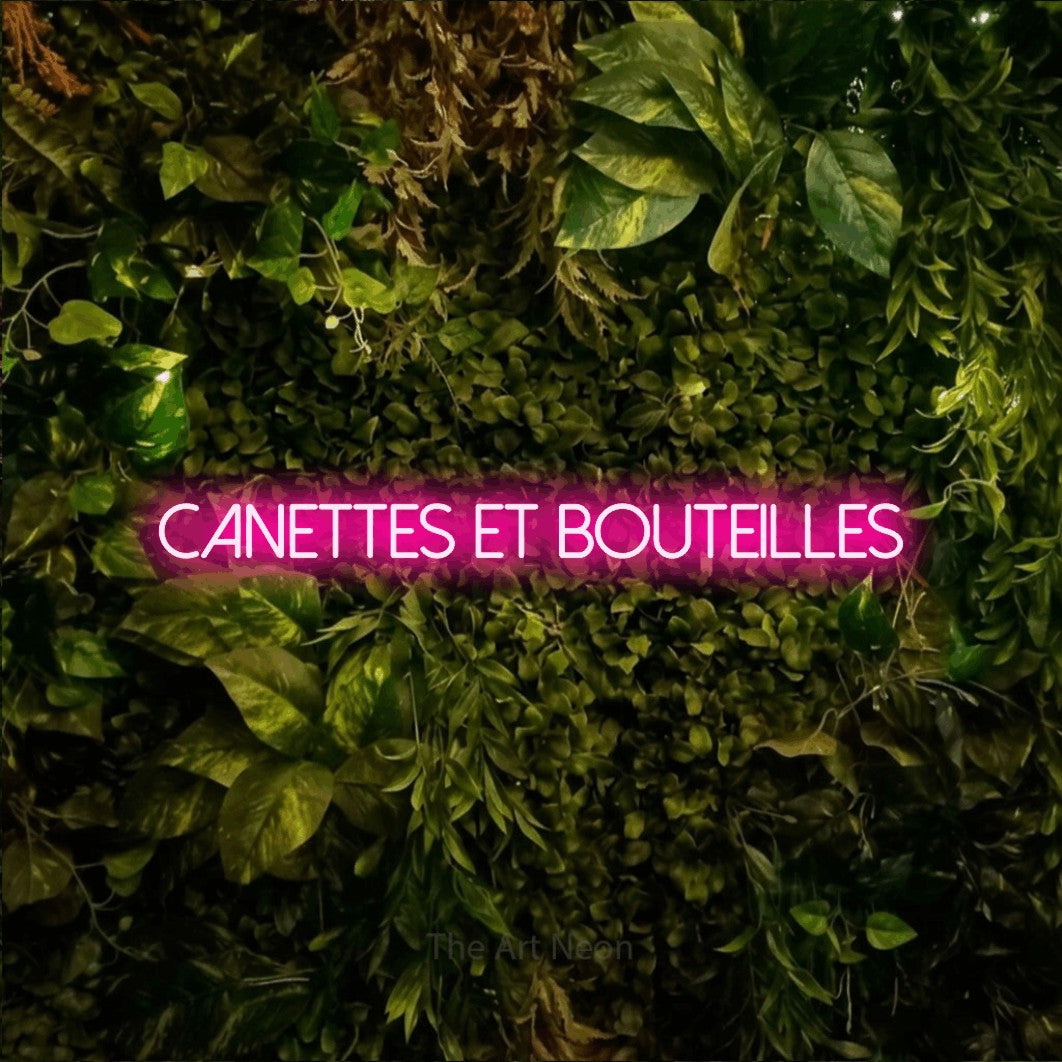 “Canettes et bouteilles” LED neon sign in hot pink, with thin sans-serif lettering, shown against bright green and yellow foliage.