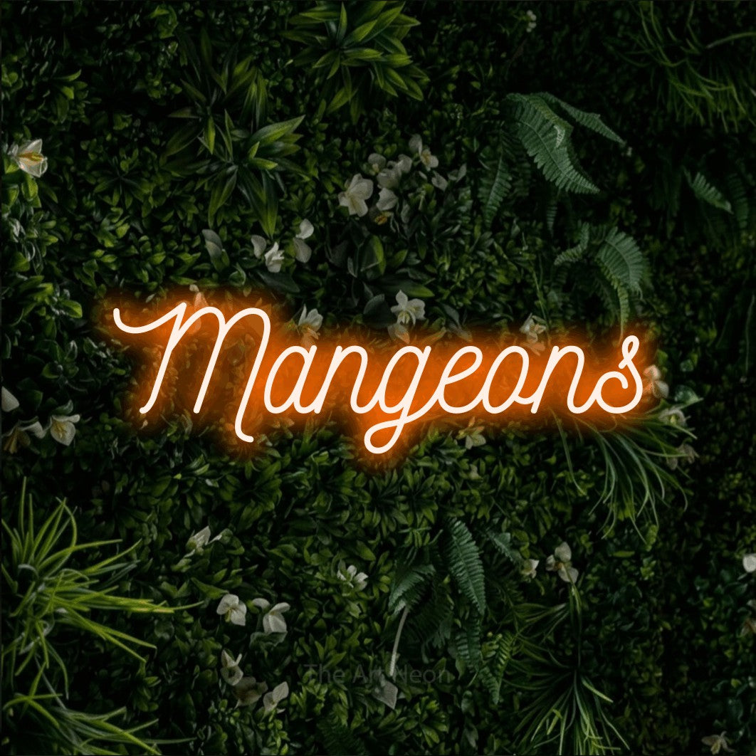 “Mangeons” LED neon sign in orange, with ornate cursive lettering, shown against a dense green plant wall.