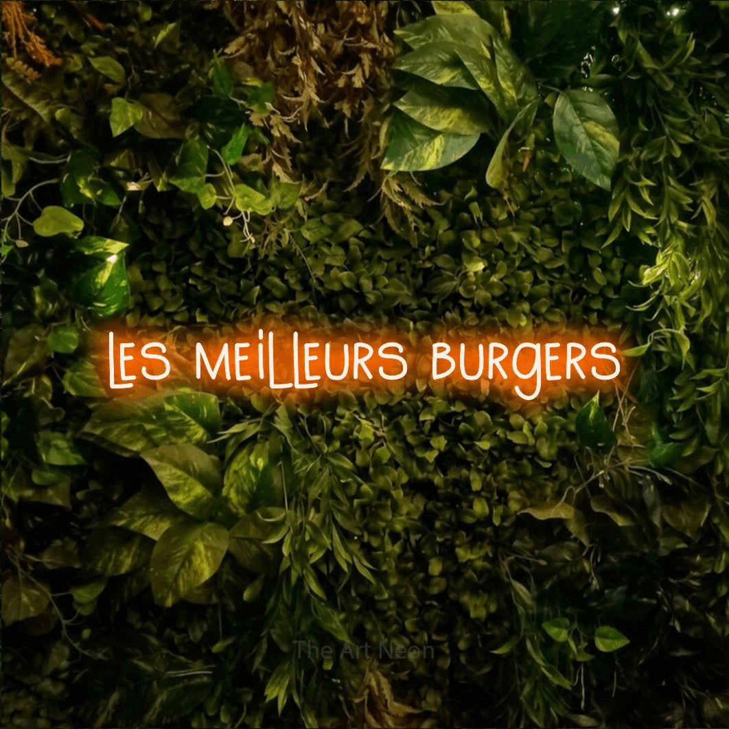 “Les meilleurs burgers” LED neon sign in orange, with narrow hand-drawn lettering, shown against bright green and yellow foliage.