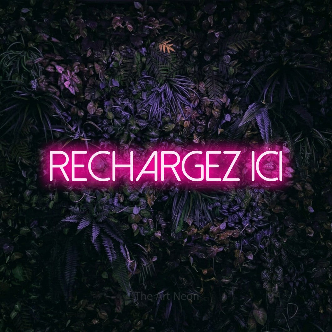 “Rechargez ici” LED neon sign in hot pink, with thin sans-serif lettering, shown against a dark wall with purple flowers.