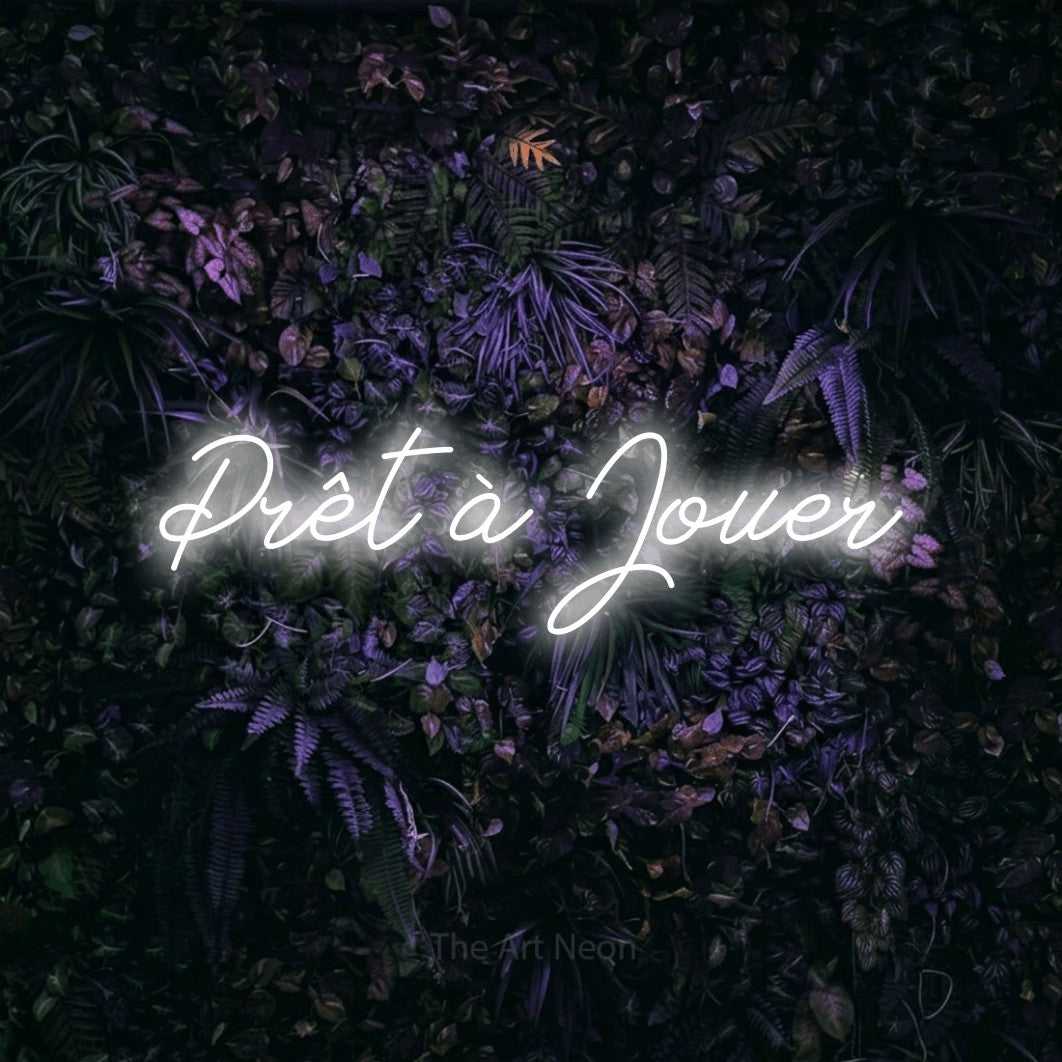 Lettrage en néon LED « Prêt à jouer » en blanc froid, en lettrage cursif, présenté sur un mur sombre aux fleurs violettes, pour un espace de jeux vidéo.