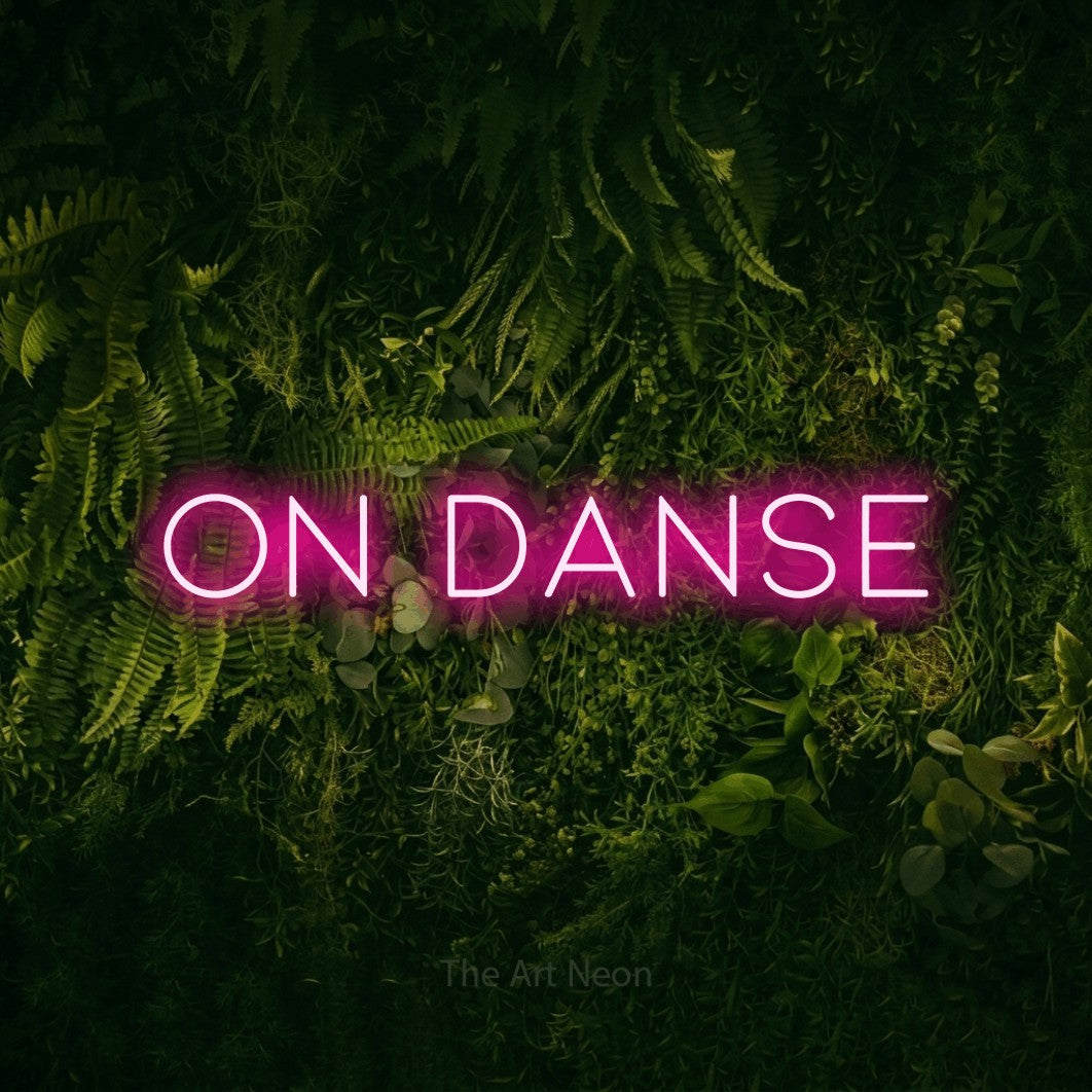 “On danse” LED neon sign in hot pink, with thin, widely spaced lettering, shown against a dark moss and forest backdrop.