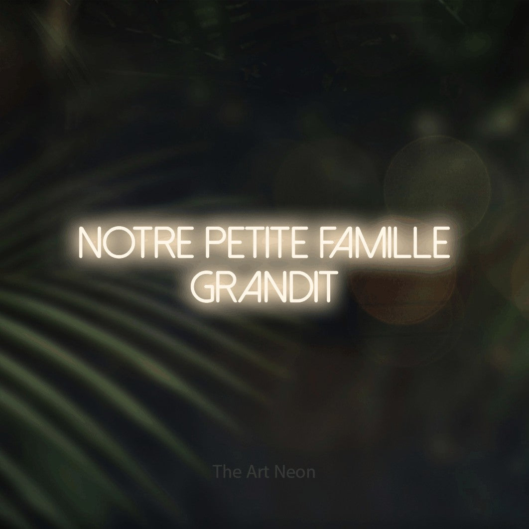 “Notre petite famille grandit” LED neon sign in warm white, with thin sans-serif lettering across 2 lines, shown against a blurred palm and bokeh-light backdrop.