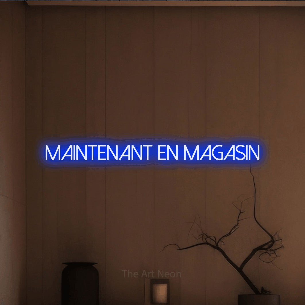 “Maintenant en magasin” LED neon sign in blue, with thin sans-serif lettering, shown against a simple earth-toned interior.