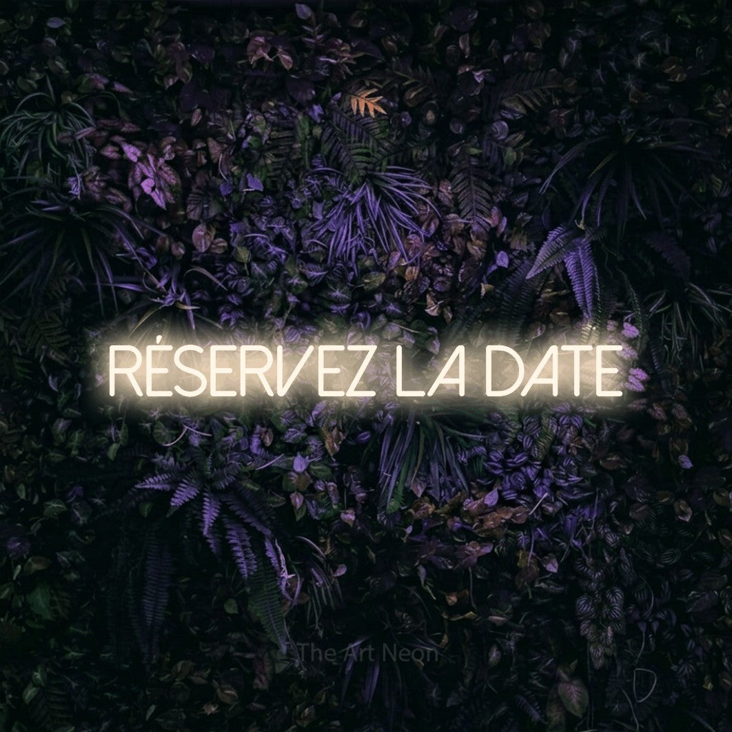 “Réservez la date” LED neon sign in warm white, with thin sans-serif lettering, shown against a dark wall with purple flowers.