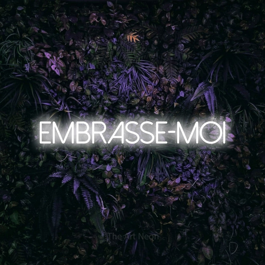 “Embrasse-moi” LED neon sign in cool white, with thin sans-serif lettering, shown against a dark wall with purple flowers.