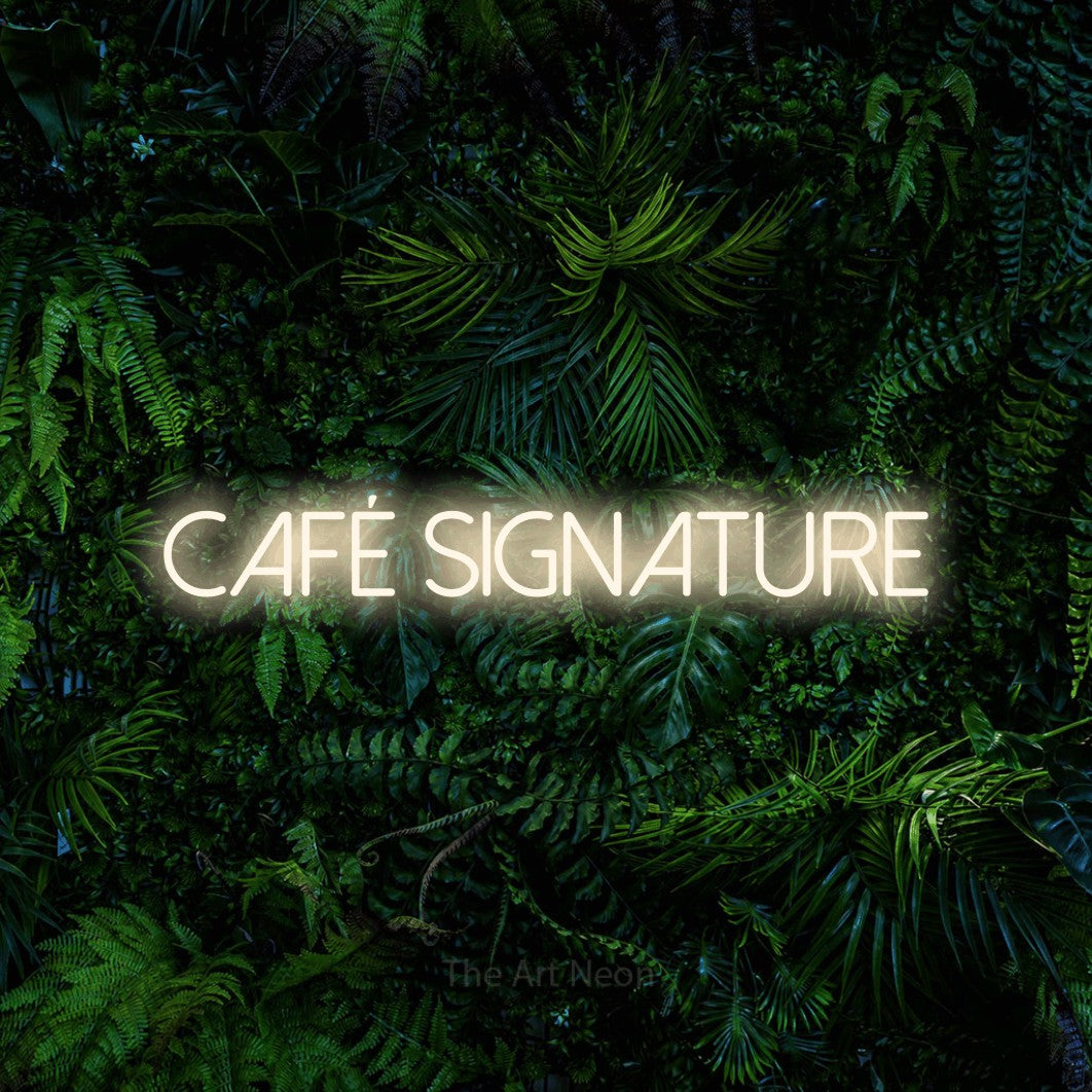 “Café signature” LED neon sign in warm white, with thin sans-serif lettering, shown against dense tropical foliage.