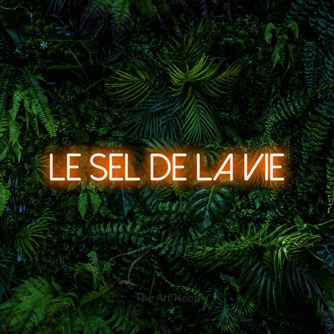 “Le sel de la vie” LED neon sign in orange, with thin sans-serif lettering, shown against dense tropical foliage.