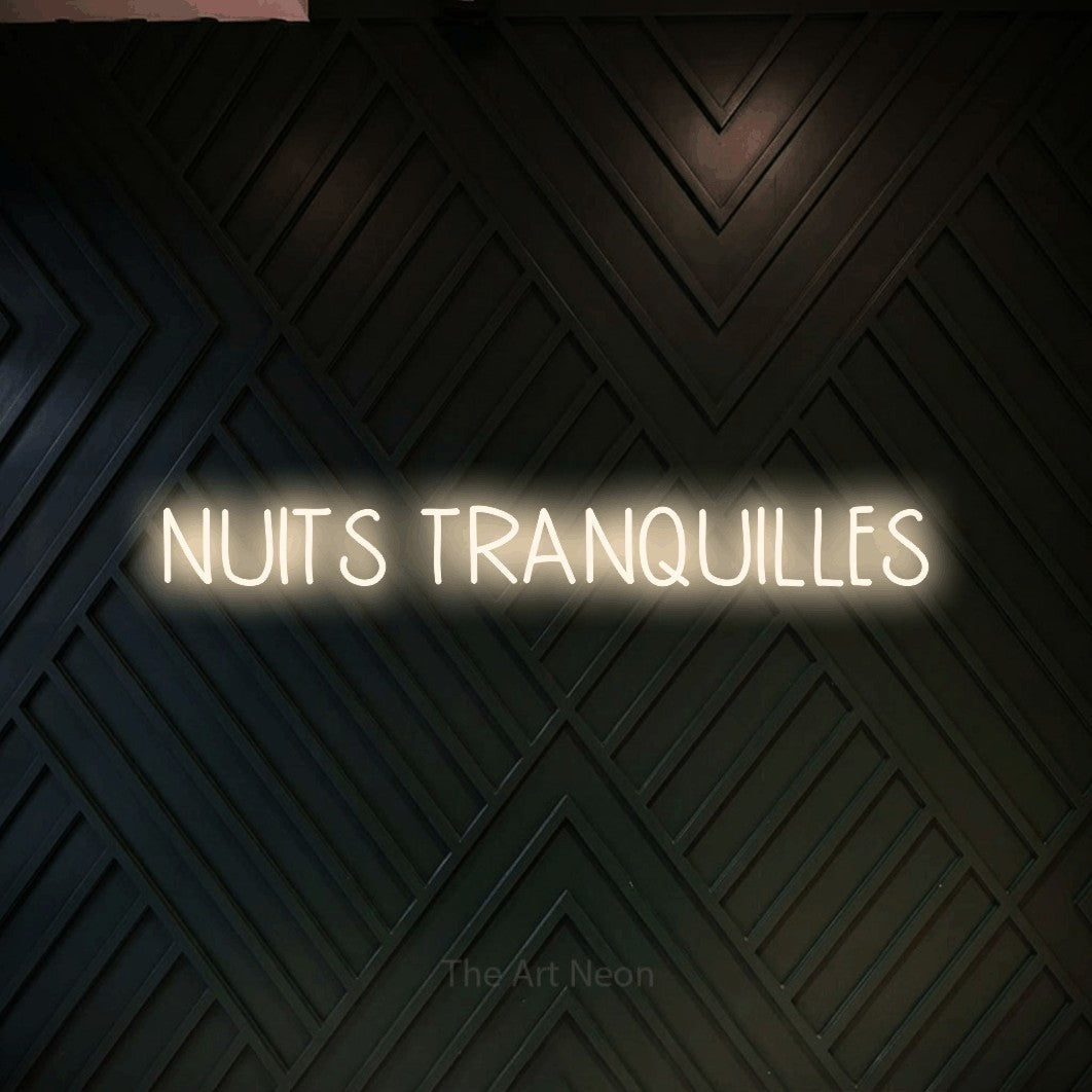“Nuits tranquilles” LED neon sign in warm white, with narrow hand-drawn lettering, shown against a black chevron-patterned wood wall.