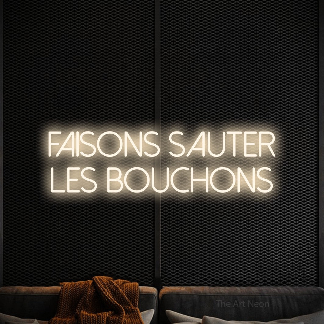 “Faisons sauter les bouchons” LED neon sign in warm white, with thin sans-serif lettering across 2 lines, shown against a modern dark-toned seating area.