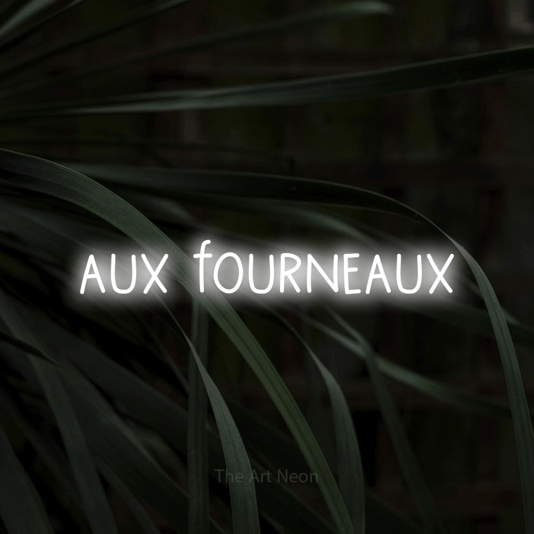 “Aux fourneaux” LED neon sign in cool white, with narrow hand-drawn lettering, shown against a close-up of dark foliage.