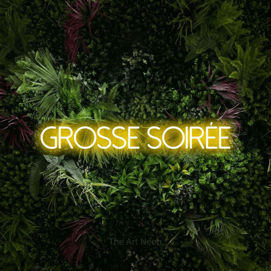 “Grosse soirée” LED neon sign in yellow, with thin sans-serif lettering, shown against a plant wall with pink flowers.