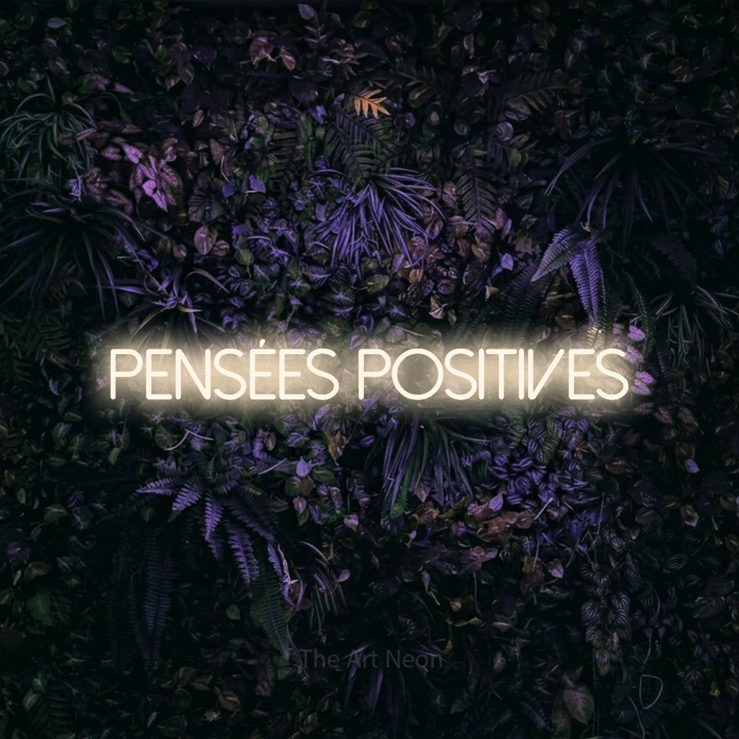“Pensées positives” LED neon sign in warm white, with thin sans-serif lettering, shown against a dark wall with purple flowers.