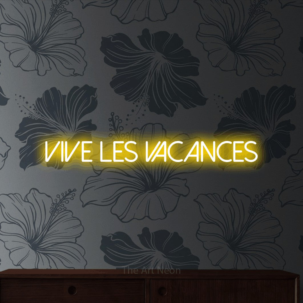 “Vive les vacances” LED neon sign in yellow, with thin sans-serif lettering, shown against a grey floral-patterned wall.