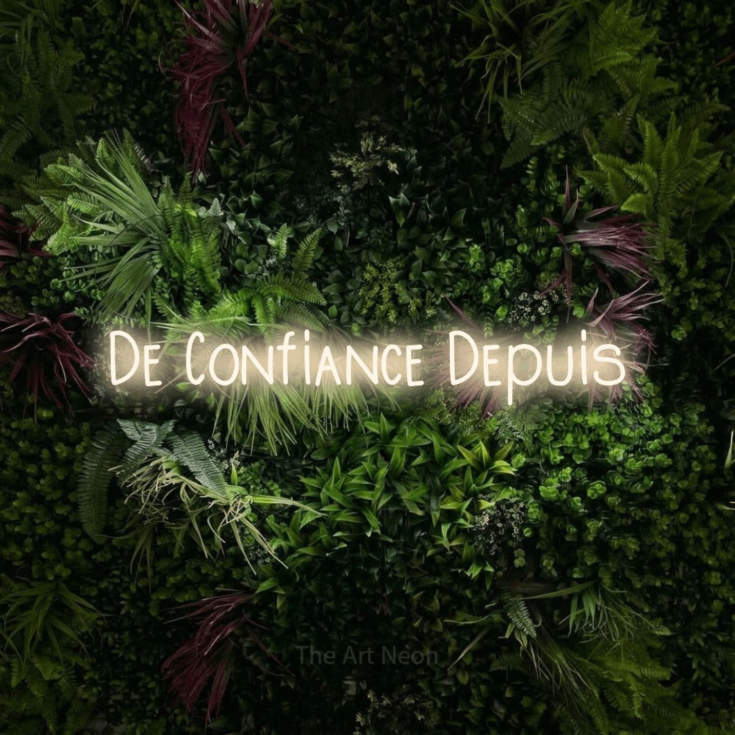 “De confiance depuis” LED neon sign in warm white, with narrow hand-drawn lettering, shown against a plant wall with pink flowers.