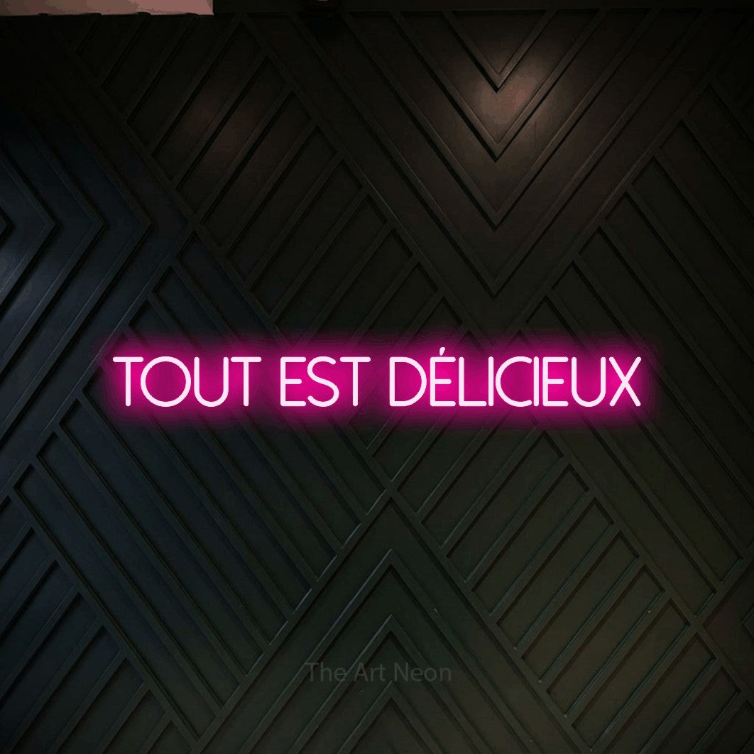“Tout est délicieux” LED neon sign in hot pink, with thin sans-serif lettering, shown against a black chevron-patterned wood wall.