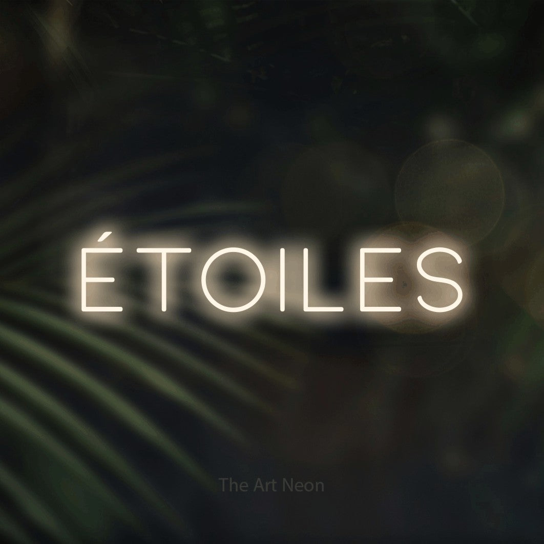 “Étoiles” LED neon sign in warm white, with thin, widely spaced lettering, shown against a blurred palm and bokeh-light backdrop.