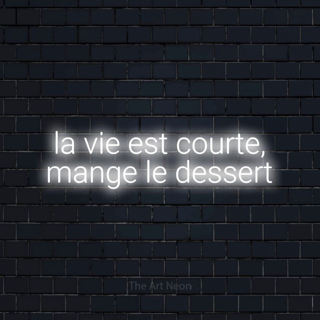 Composition en néon LED « La vie est courte, mange le dessert » sur deux lignes en blanc froid, en lettres sans empattement arrondies, mise en valeur sur un mur de briques bleu marine foncé, pour une cuisine.