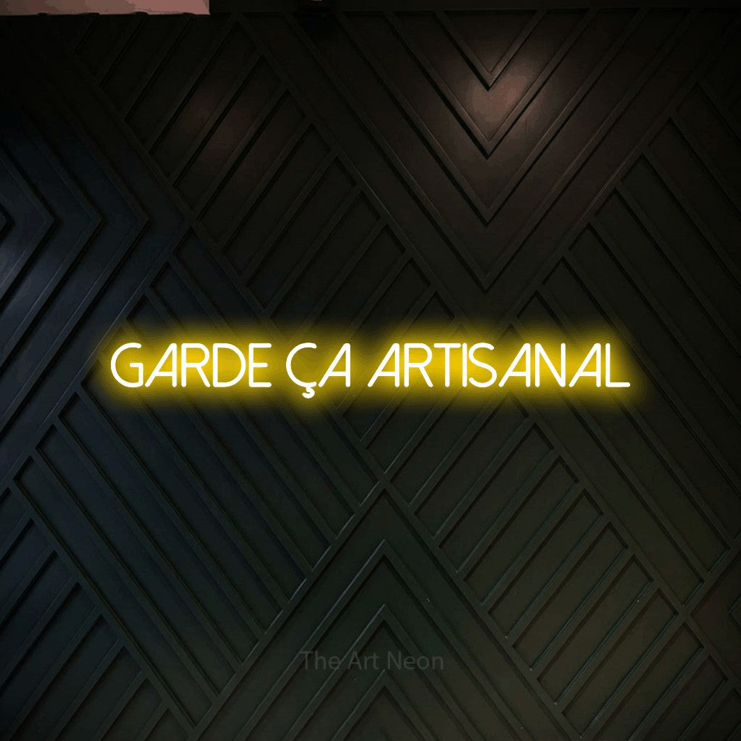 “Garde ça artisanal” LED neon sign in yellow, with thin sans-serif lettering, shown against a black chevron-patterned wood wall.