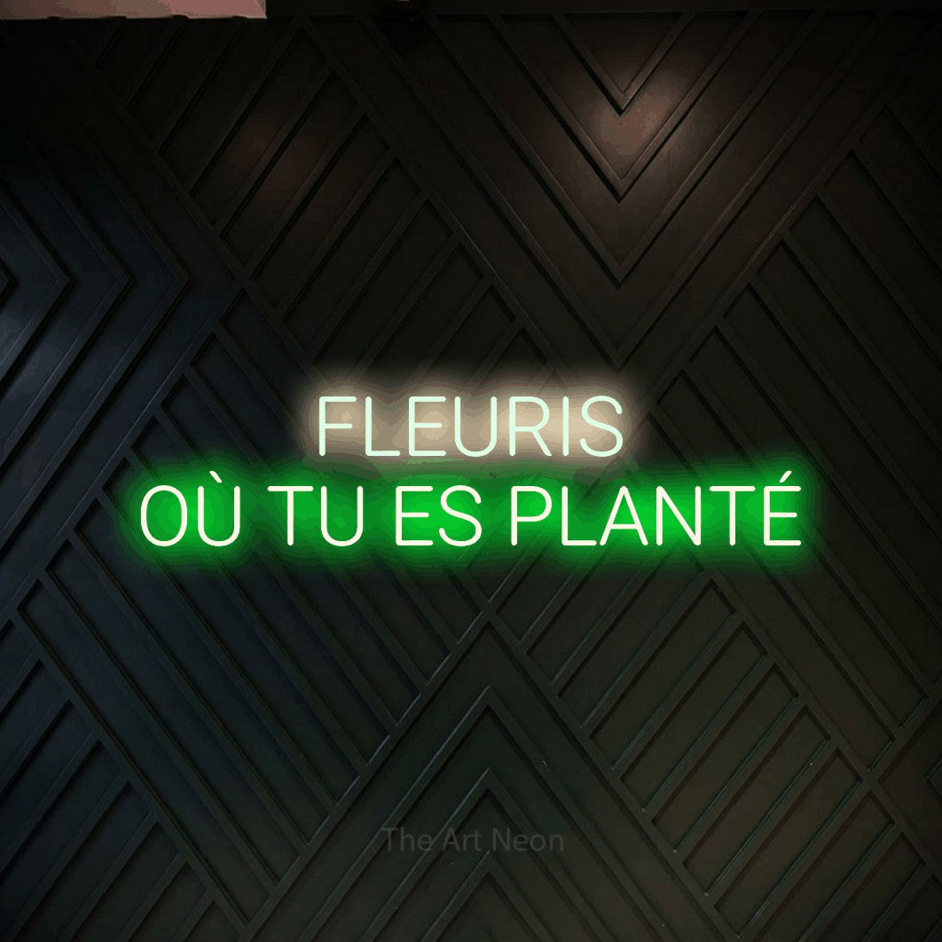 “Fleuris où tu es planté” LED neon sign in warm white on the upper line and green on the lower line, with rounded sans-serif lettering across 2 lines, shown against a black chevron-patterned wood wall.