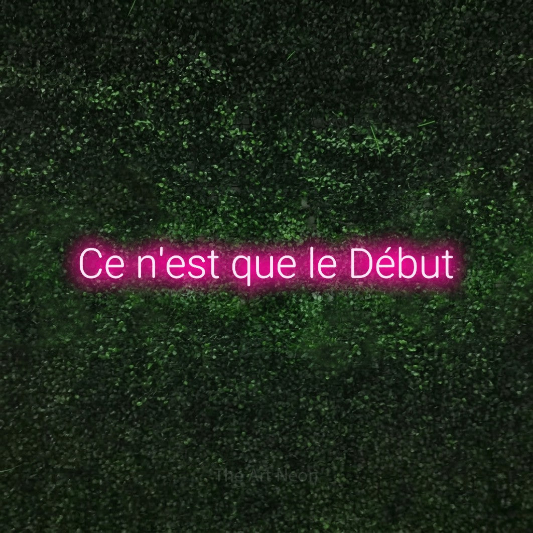 “Ce n'est que le début” LED neon sign in hot pink, with rounded sans-serif lettering, shown against a textured green hedge.