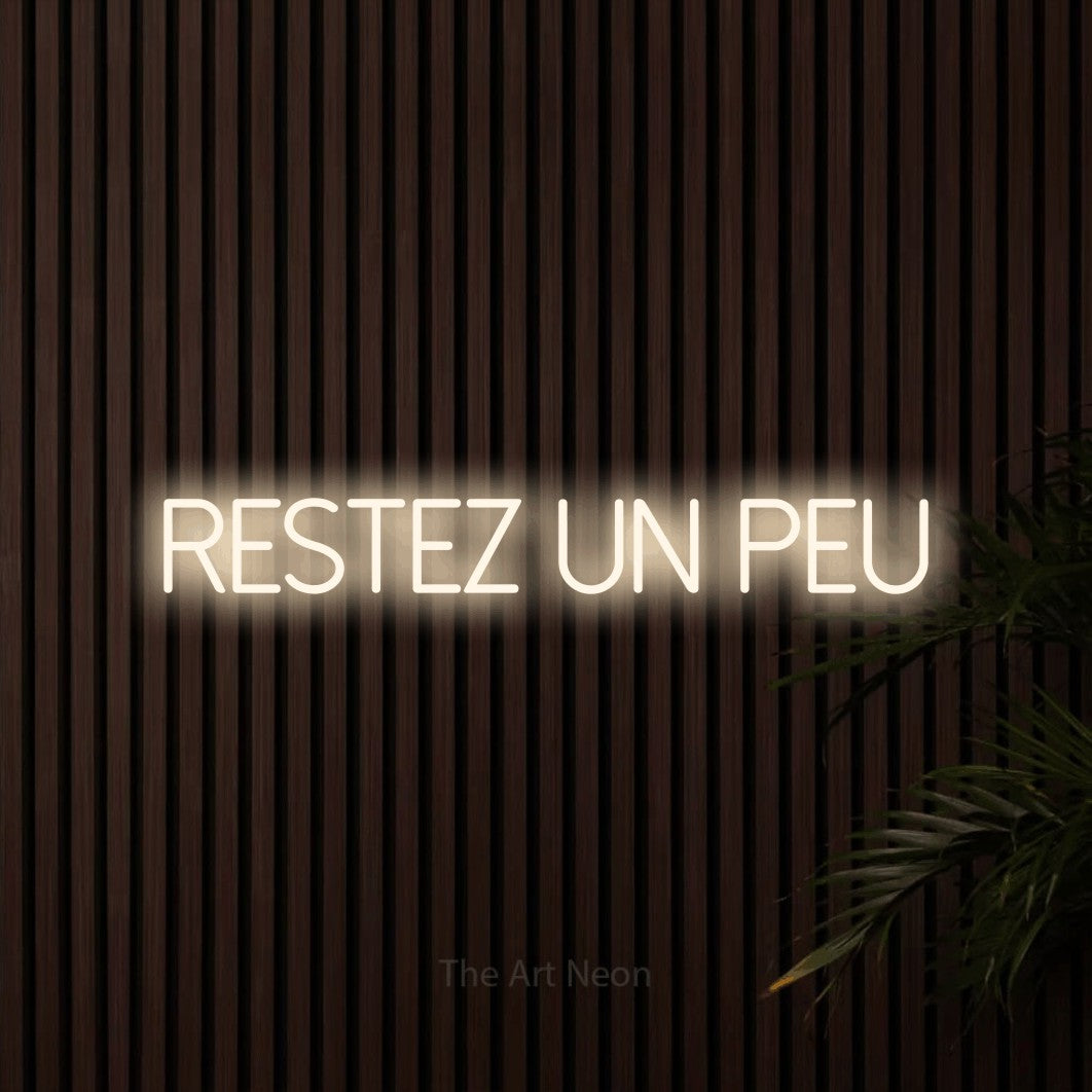 Néon LED « Restez un peu » en blanc chaud, en lettres fines sans empattement, mis en scène sur un mur de lattes en bois foncé, pour un salon.