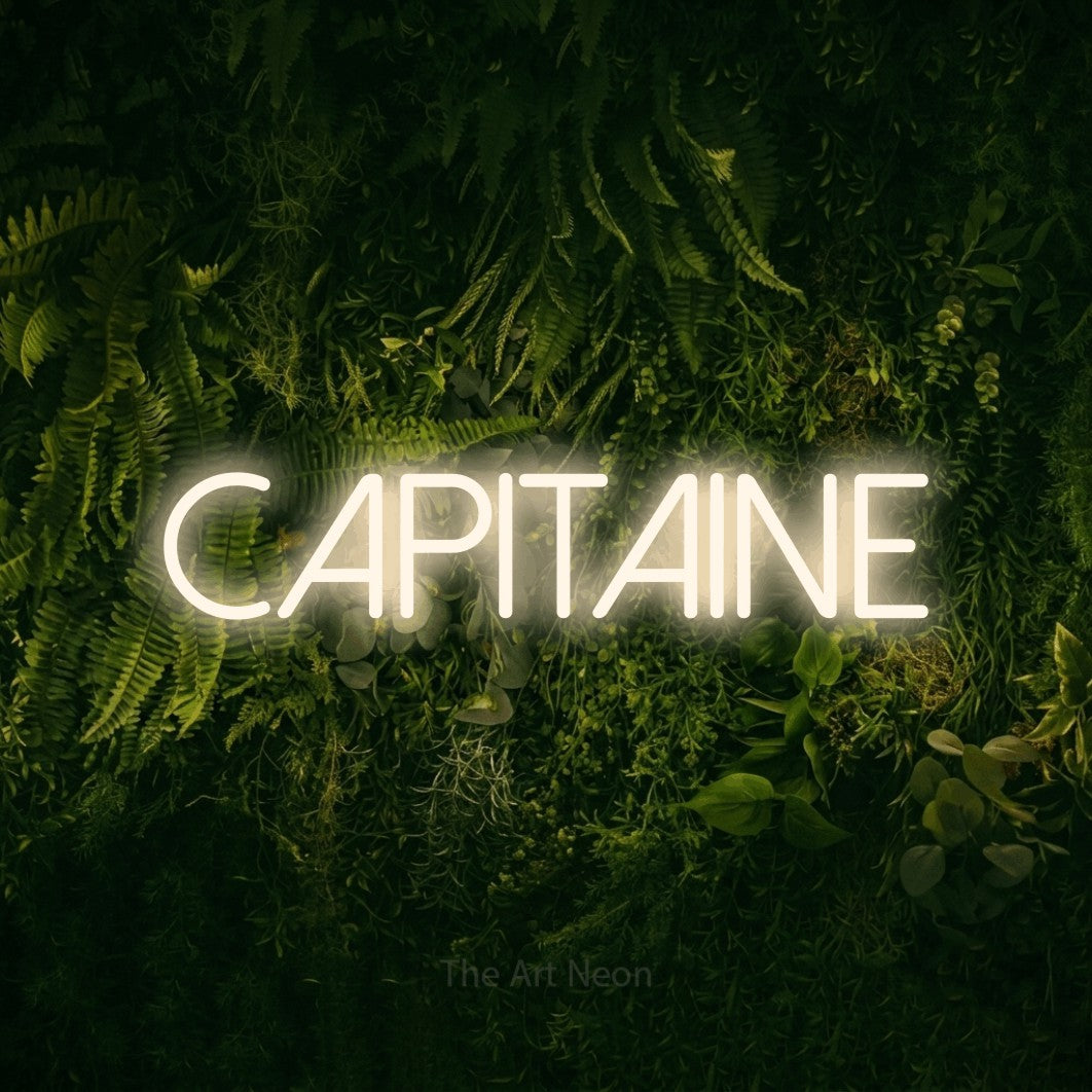 “Capitaine” LED neon sign in warm white, with thin sans-serif lettering, shown against a dark moss and forest backdrop.