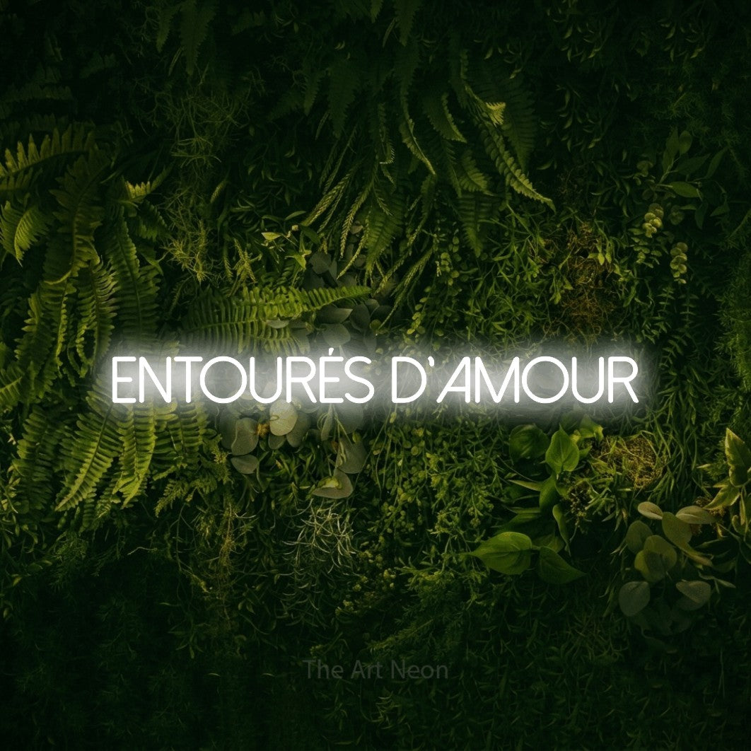 “Entourés d'amour” LED neon sign in cool white, with thin sans-serif lettering, shown against a dark moss and forest backdrop.