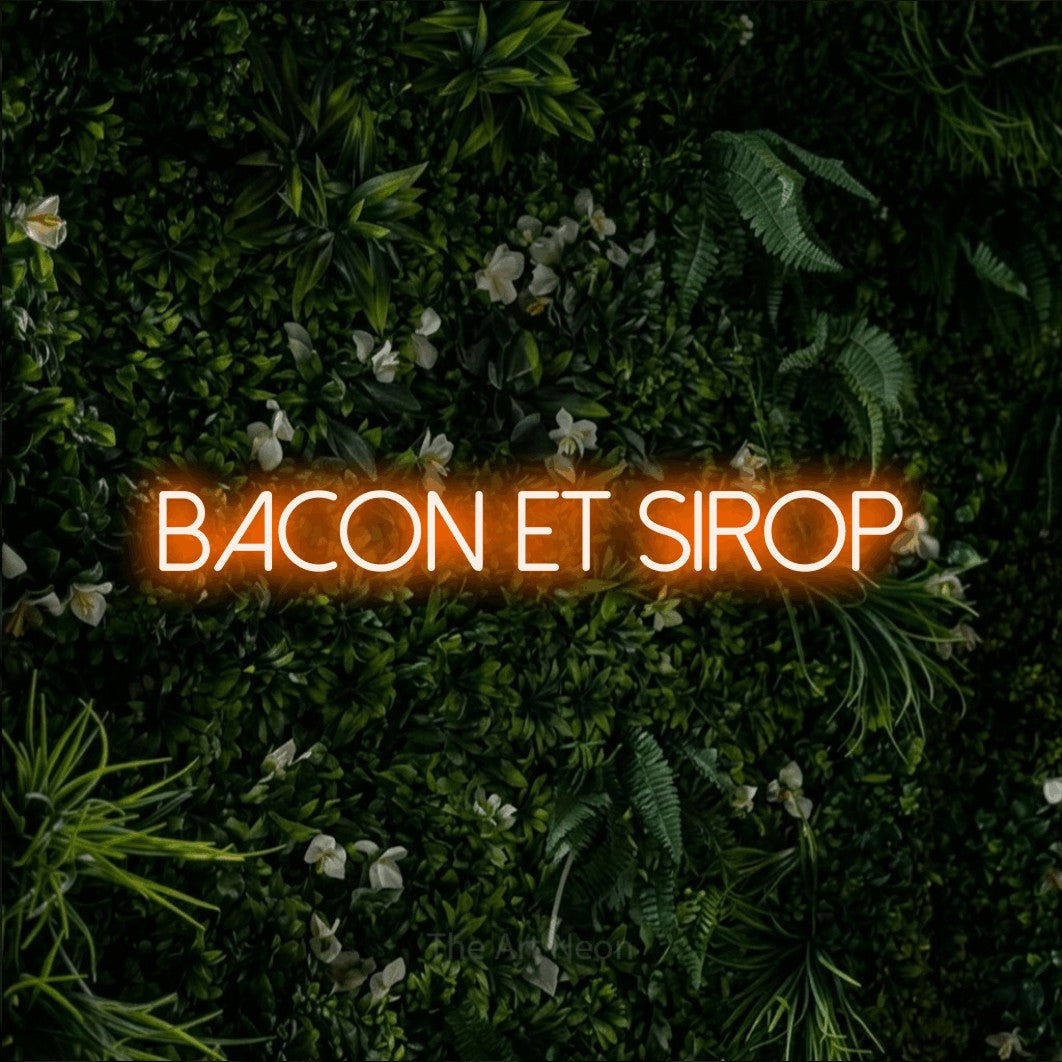 “Bacon et sirop” LED neon sign in orange, with thin sans-serif lettering, shown against a dense green plant wall.