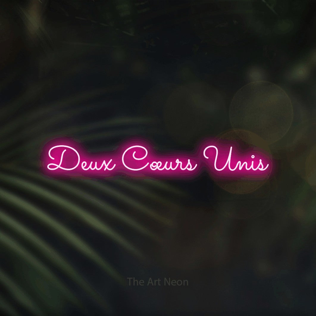 “Deux cœurs unis” LED neon sign in hot pink, with looped cursive lettering, shown against a blurred palm and bokeh-light backdrop.