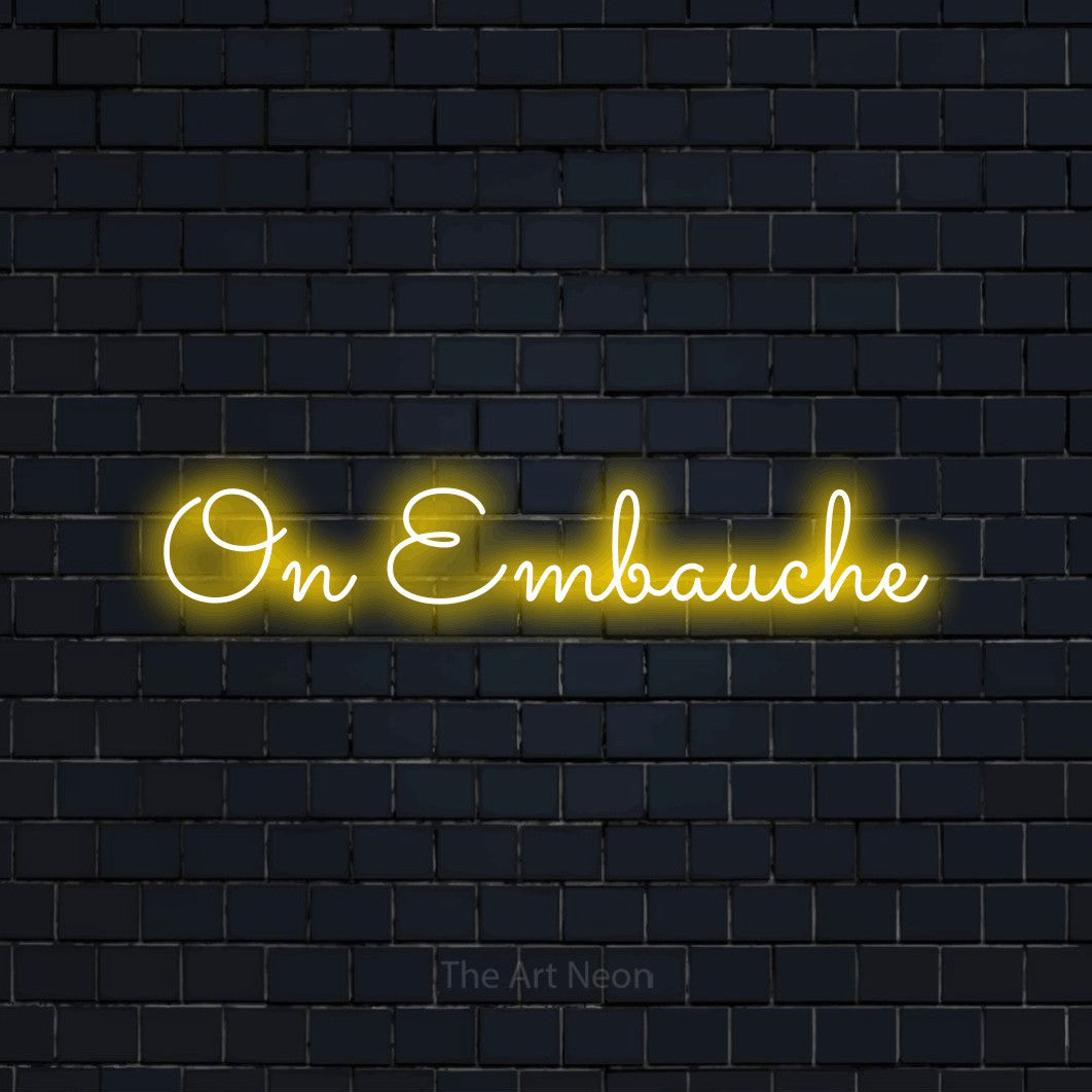 “On embauche” LED neon sign in yellow, with looped cursive lettering, shown against a dark navy brick wall.