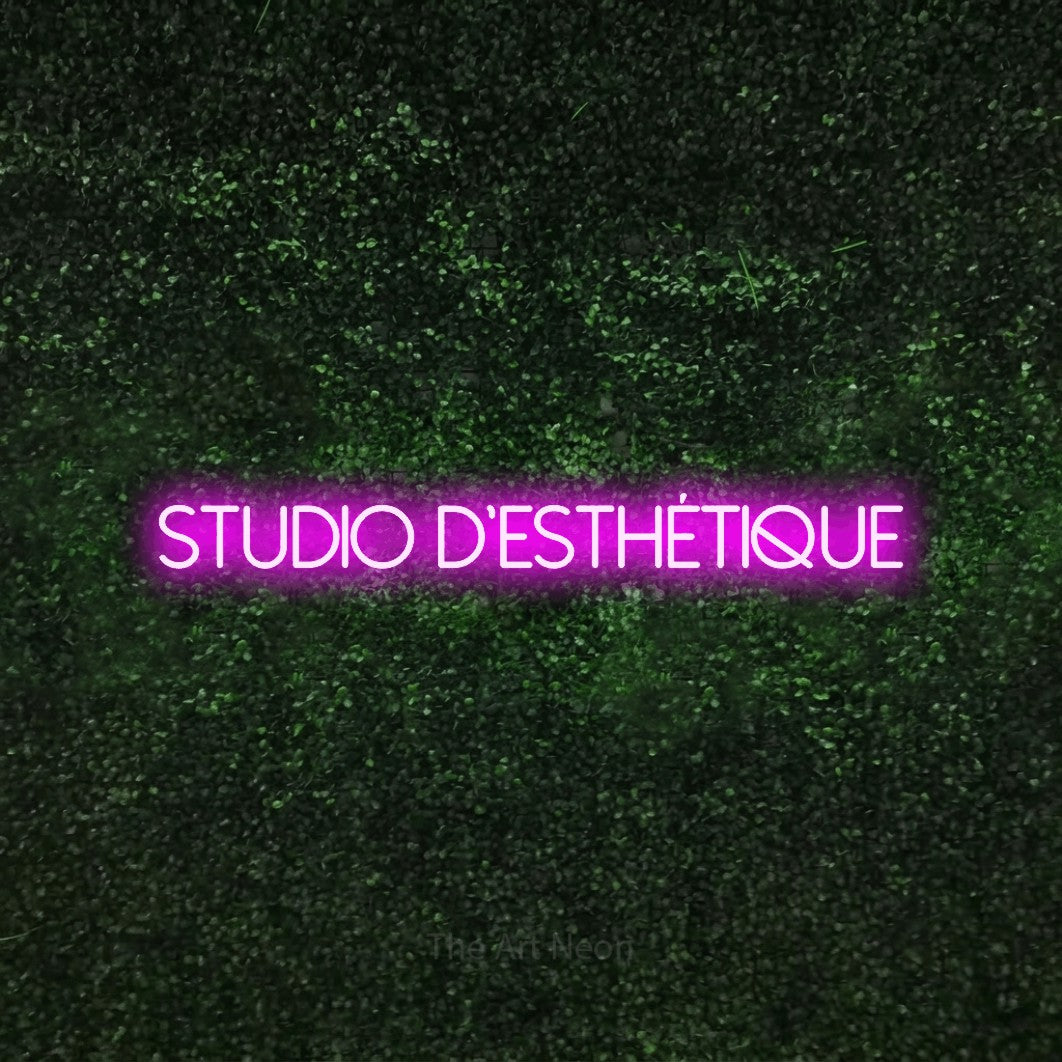 Décoration murale en néon LED « Studio d'esthétique » en rose, en lettres fines sans empattement, installée sur une haie verte texturée, pour un salon de beauté.