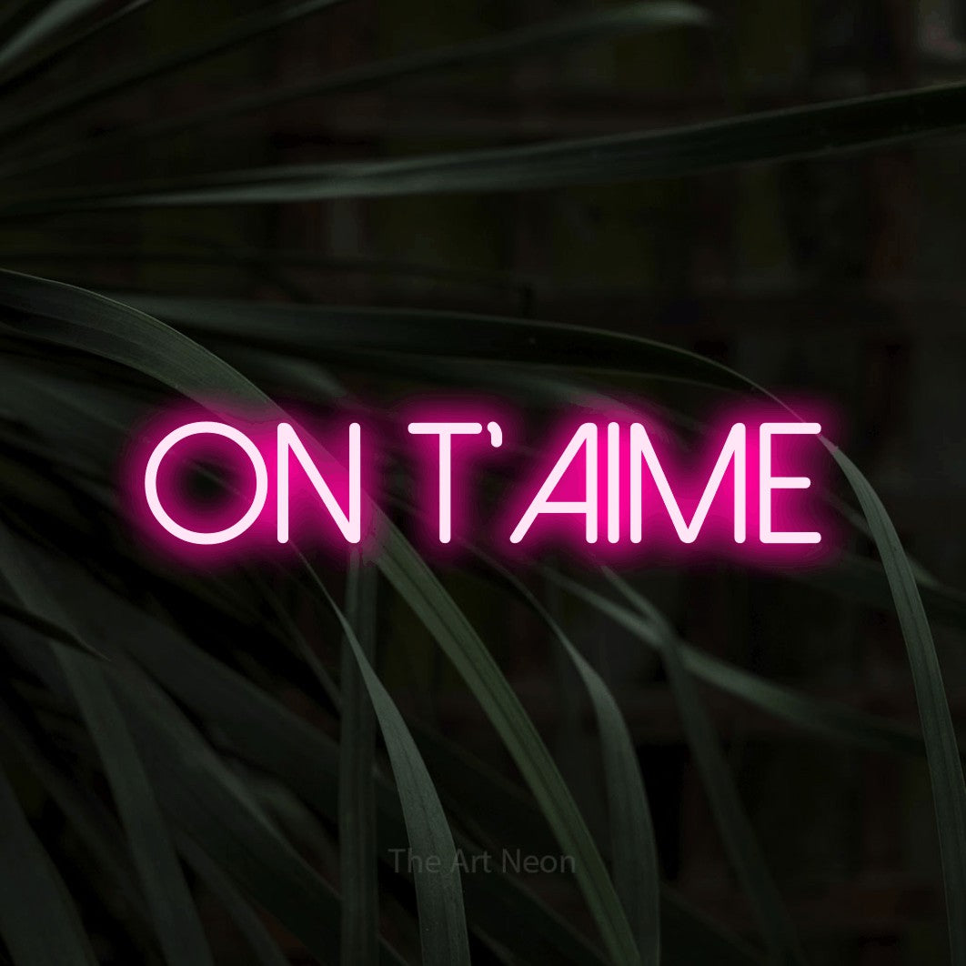 “On t'aime” LED neon sign in hot pink, with thin sans-serif lettering, shown against a close-up of dark foliage.
