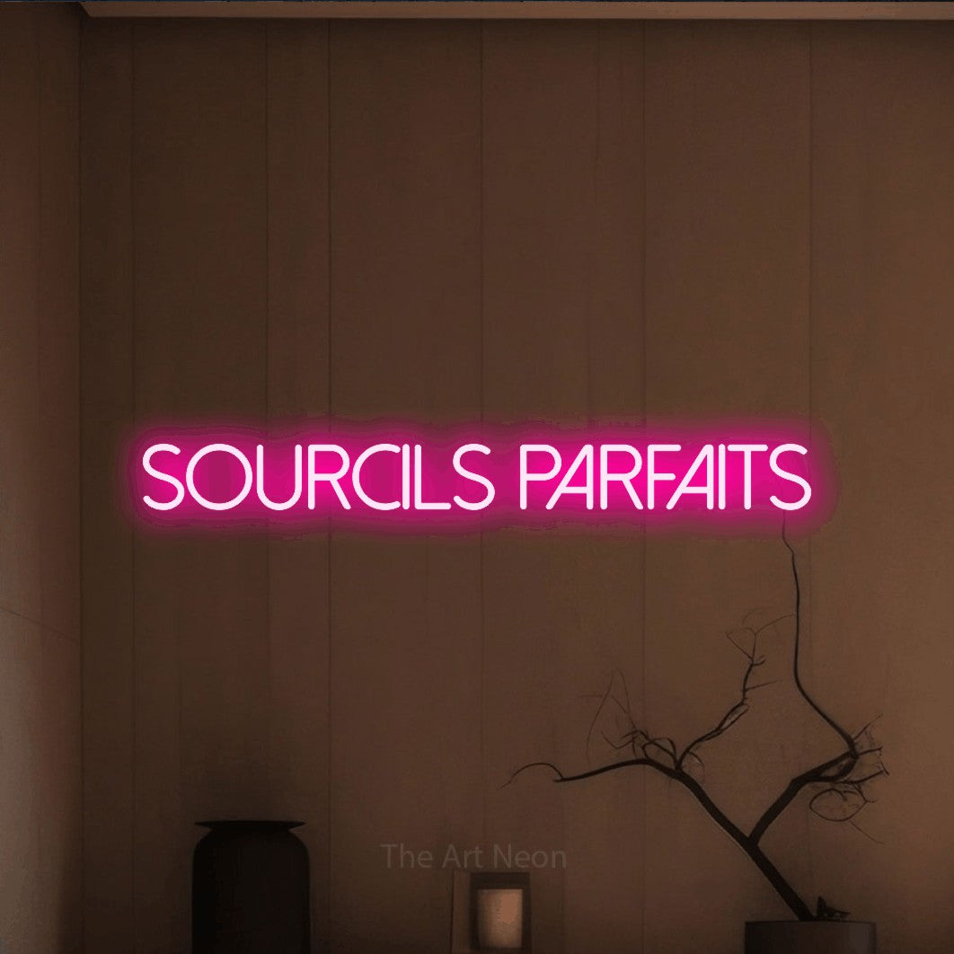 “Sourcils parfaits” LED neon sign in hot pink, with thin sans-serif lettering, shown against a simple earth-toned interior.