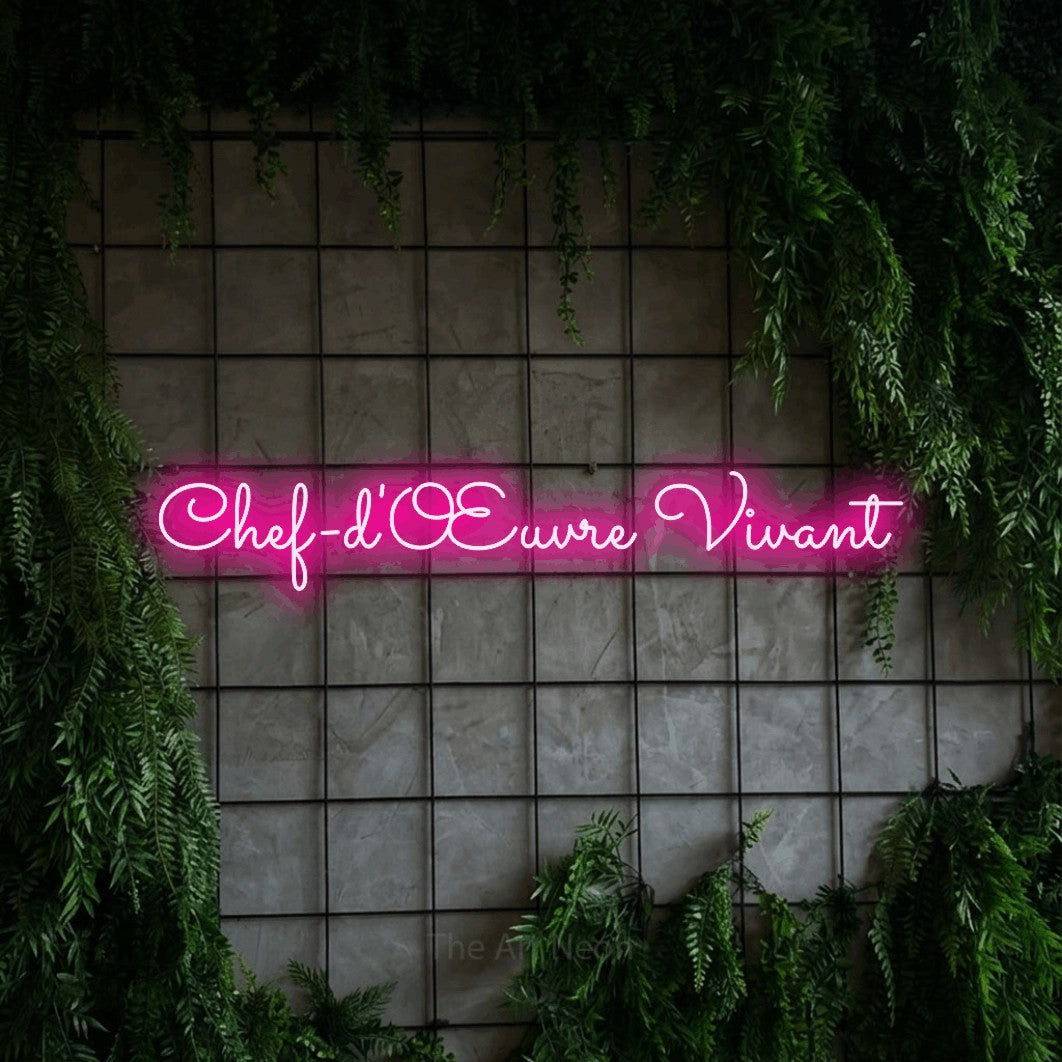 “Chef-d'œuvre vivant” LED neon sign in hot pink, with looped cursive lettering, shown against an ivy-covered concrete grid.
