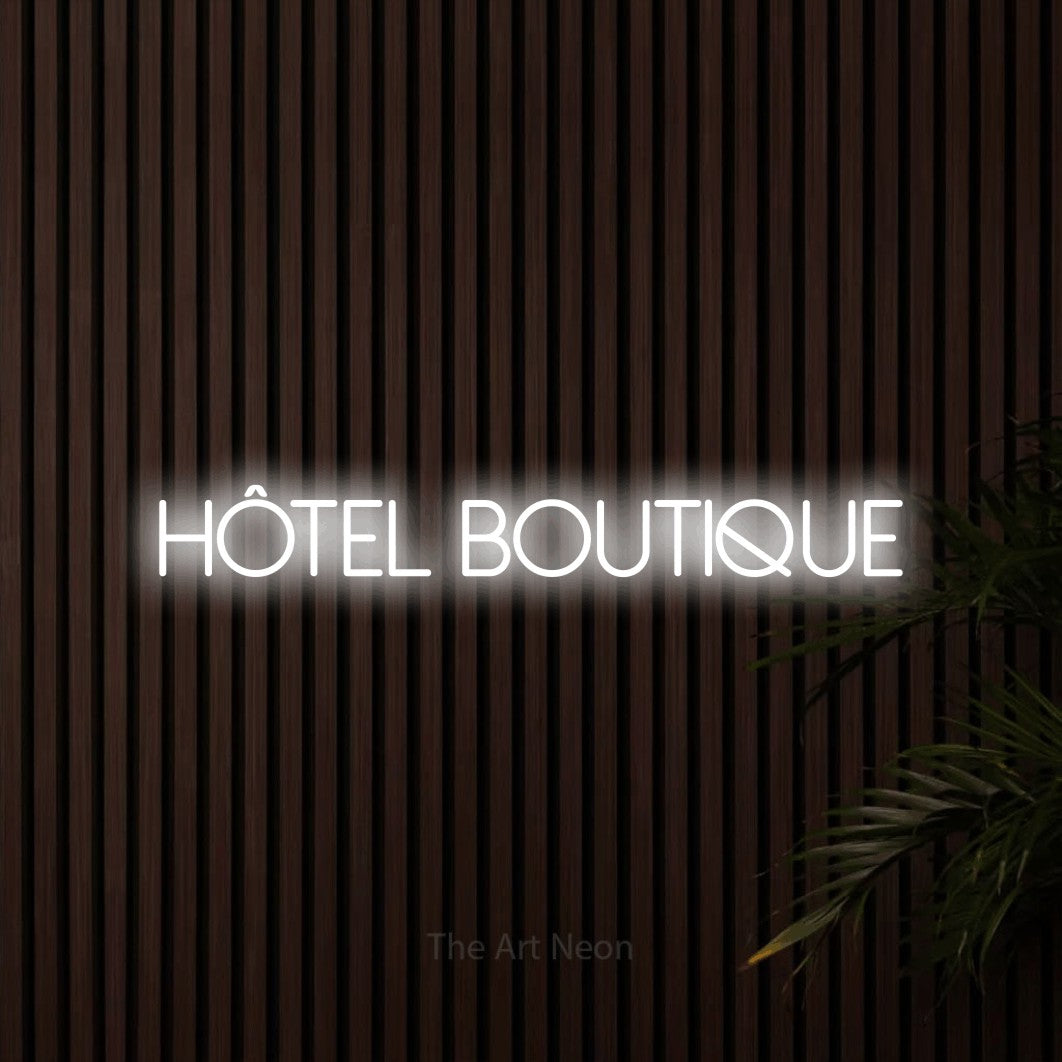 Lettrage en néon LED « Hôtel boutique » en blanc froid, en lettres fines sans empattement, présenté sur un mur de lattes en bois foncé, pour un hôtel.