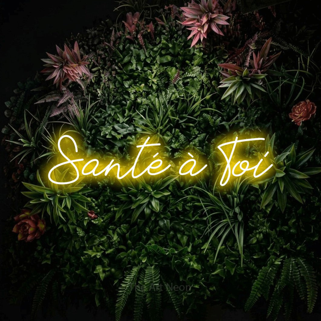 “Santé à toi” LED neon sign in yellow, with cursive lettering, shown against a flowering plant wall.