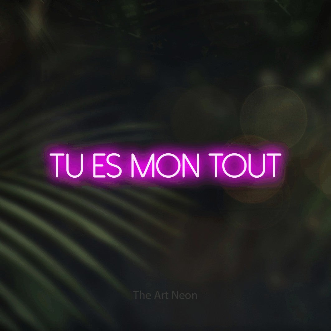 “Tu es mon tout” LED neon sign in pink, with thin sans-serif lettering, shown against a blurred palm and bokeh-light backdrop.