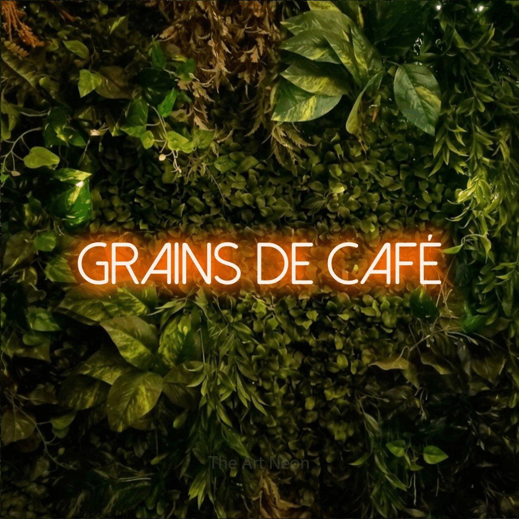 “Grains de café” LED neon sign in orange, with thin sans-serif lettering, shown against bright green and yellow foliage.