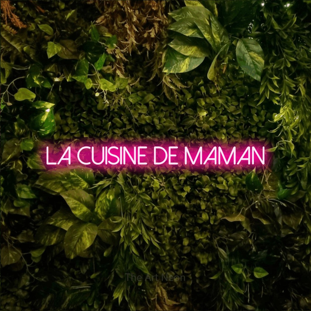“La cuisine de maman” LED neon sign in hot pink, with thin sans-serif lettering, shown against bright green and yellow foliage.