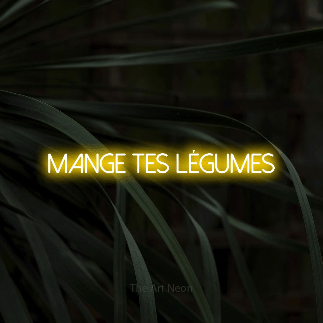 “Mange tes légumes” LED neon sign in yellow, with thin sans-serif lettering, shown against a close-up of dark foliage.